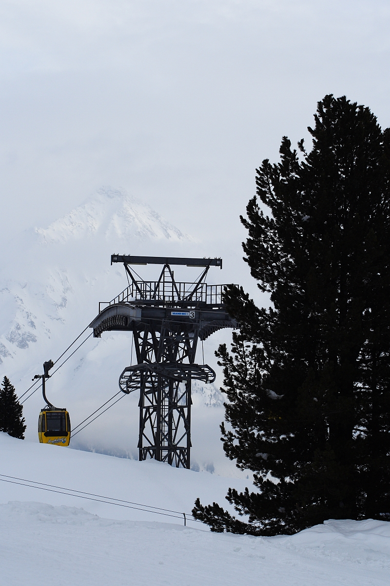 A yellow cable car ascends on a cable towards a metal support tower in the Austrian Alps. The scene is set in a snowy mountainous area with snow-covered peaks visible in the background. To the right of the tower, tall, dark evergreens provide contrast to the white landscape.