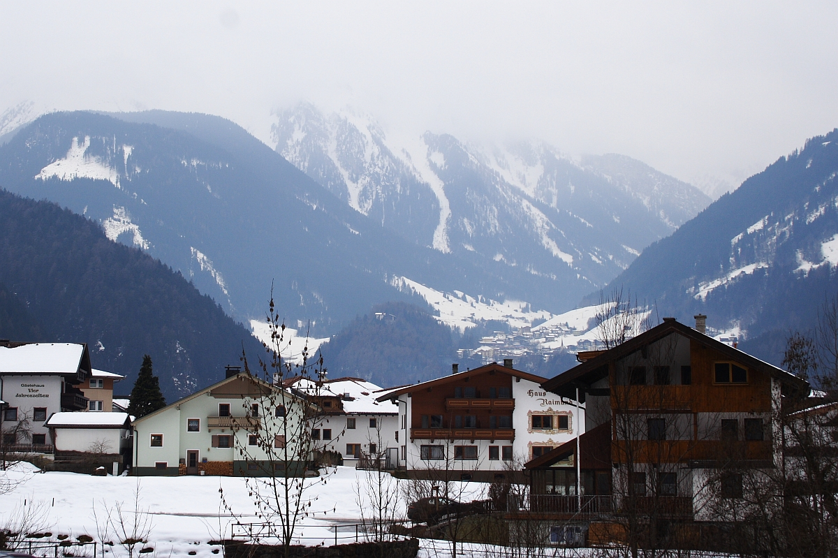 A serene, snow-covered village nestled in a mountainous region of the Austrian Alps is depicted. Traditional wooden houses with white facades are scattered amidst bare trees. The towering mountains in the background are partly shrouded in mist, and patches of snow can be seen on their slopes. The scene is tranquil and picturesque.