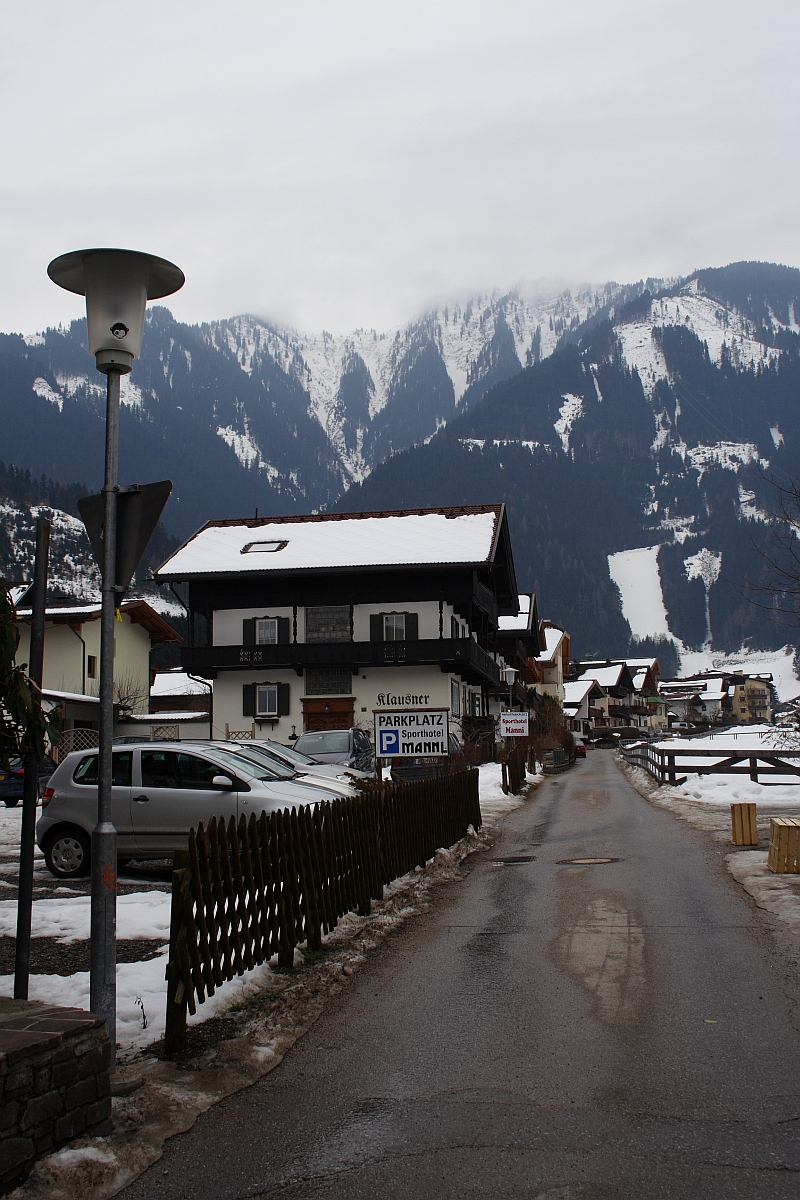 A narrow asphalt road lined with wooden fences runs through a snow-covered village in the Austrian Alps. Traditional alpine buildings with dark wooden roofs sit beside the road. A snow-capped mountain range is visible in the background under an overcast sky. A streetlamp and parked cars are on the left.