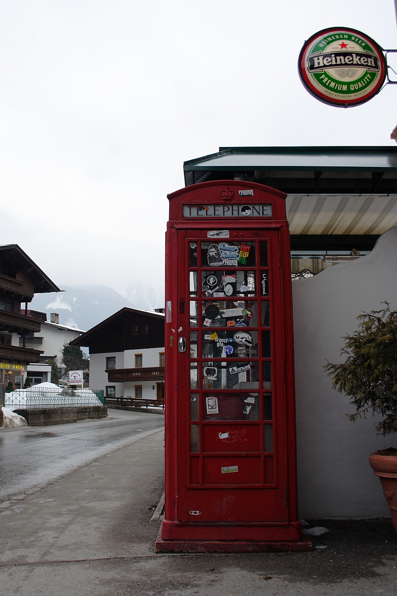 A traditional red British telephone booth stands on a wet street in a small town with alpine-style buildings, reminiscent of an Austrian village. The booth is adorned with stickers. In the upper right corner, a round Heineken beer sign is attached to a building. Snowy Alps are visible in the background.