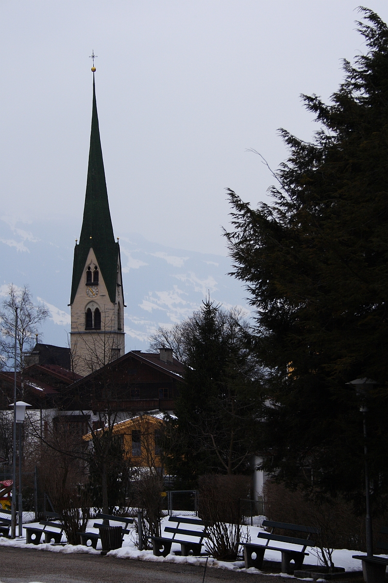 A tall church steeple with a green roof stands prominently in a snow-covered town, reminiscent of the Austrian Alps. Surrounding buildings are nestled among leafless trees, and several benches are visible in the foreground. The sky is overcast, adding a somber tone to the winter scene.