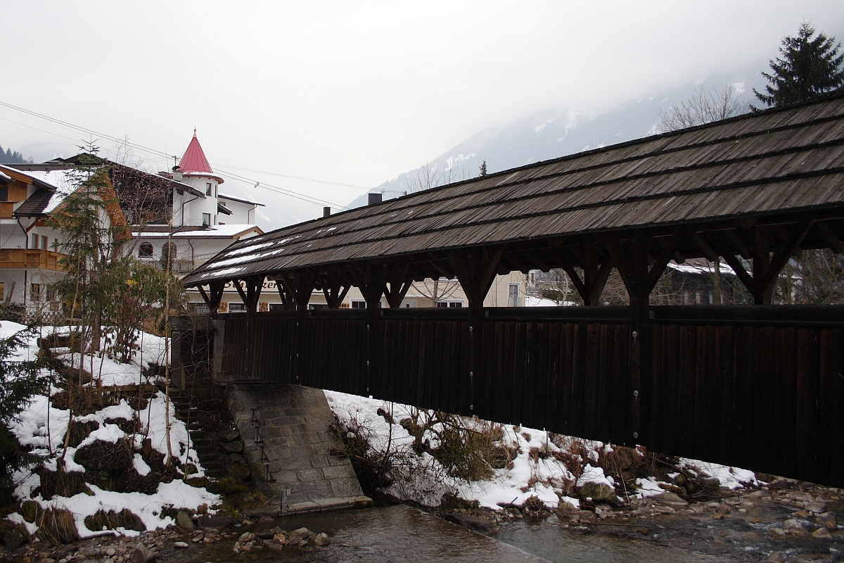 A covered wooden pedestrian bridge spans over a narrow stream in a snowy landscape. A stone path leads to the bridge, and snow patches are visible on the ground and rooftops. In the background, a church with a red steeple stands among buildings and trees, set against the misty Austrian Alps.