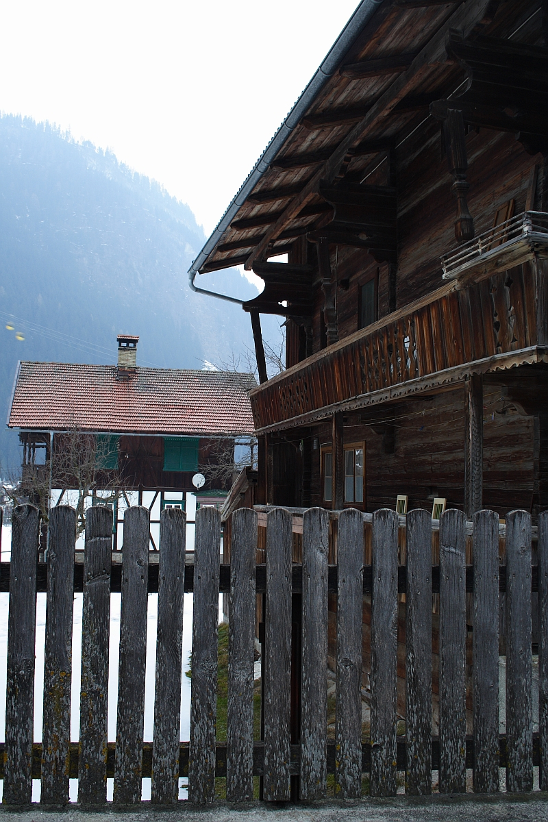 A rustic wooden house with a slanted roof and a balcony is set against the majestic Austrian Alps. A weathered wooden fence is in the foreground, and a second wooden building with a chimney is visible behind. The scene appears serene and misty, suggesting a cold, possibly winter day in Austria.