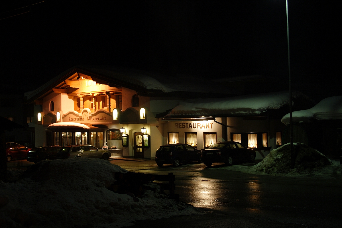 A cozy restaurant named "Mittergrat" in the snow-covered Austrian Alps is illuminated at night. The building features large windows and warm lights, casting a soft glow on the snowy ground and parked cars. A streetlight provides additional lighting, and snow piles can be seen in the foreground.