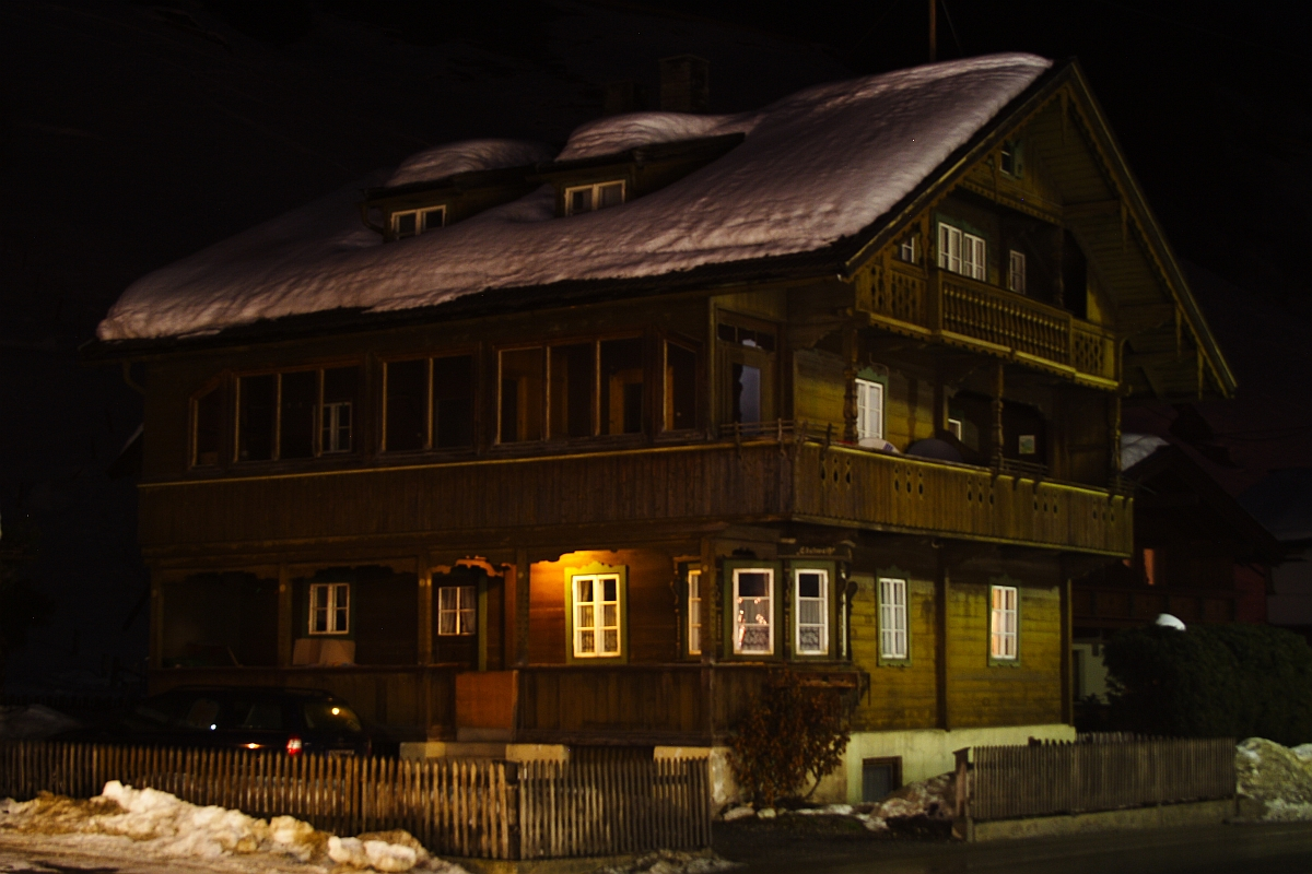 A wooden alpine house with a snow-covered roof at night, nestled in the Austrian Alps. Warm yellow lights glow through some windows. The house has a balcony with ornate wooden railings. A fence surrounds the property; a car is partially visible on the left side near the entrance amidst the snow-covered ground.