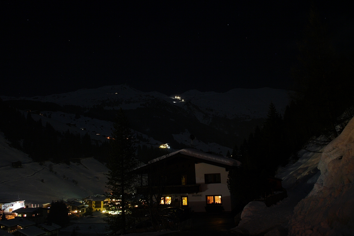 A serene nighttime mountain village scene in the Austrian Alps. Dim lights from houses illuminate the snowy landscape. A dark wooden chalet is in the foreground with warm lights in the windows. Snow-covered slopes and tall trees are set against a starry sky, with distant peaks barely visible in the dark.
