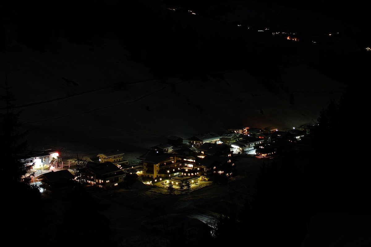 A nighttime view of a small, illuminated village nestled in the Austrian Alps. The buildings are lit with warm, yellow lights, creating a cozy atmosphere amidst the surrounding darkness. Snow-covered hills and trees are faintly visible in the background, hinting at a winter setting.
