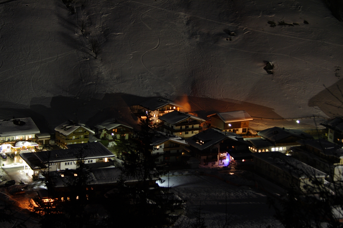 A nighttime aerial view of a snowy alpine village in the Austrian Alps, illuminated by warm yellow lights from the windows and street lamps. The snow-covered rooftops and surrounding snow-covered landscape create a serene, wintry atmosphere. Tracks in the snow are visible in the background on the hillside.