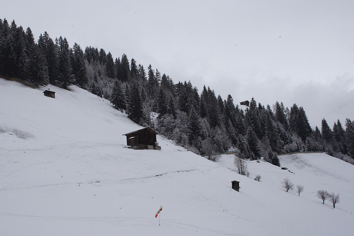 A snowy mountain landscape in the Austrian Alps, with a few small wooden huts scattered on a hillside. The hillside is covered in snow with sparse evergreen trees on the upper half. A windsock is visible near the bottom of the image. The overcast sky adds to the serene and quiet atmosphere.