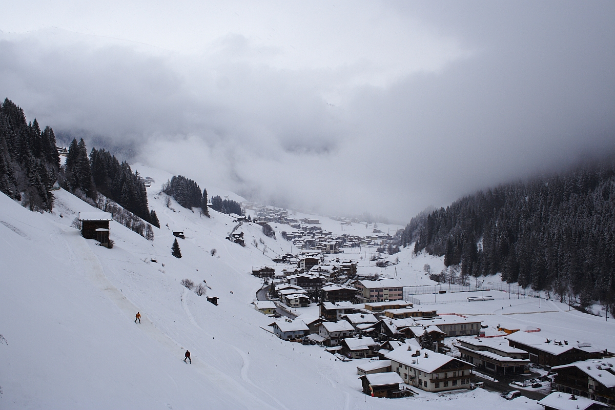 A snow-covered village nestled in a valley surrounded by the dense, evergreen forests and mountains of the Austrian Alps. The village features numerous buildings with snow-capped roofs. Skiers are visible on the slopes, while mist and clouds partly obscure the mountains in the background, adding a serene, wintry atmosphere.