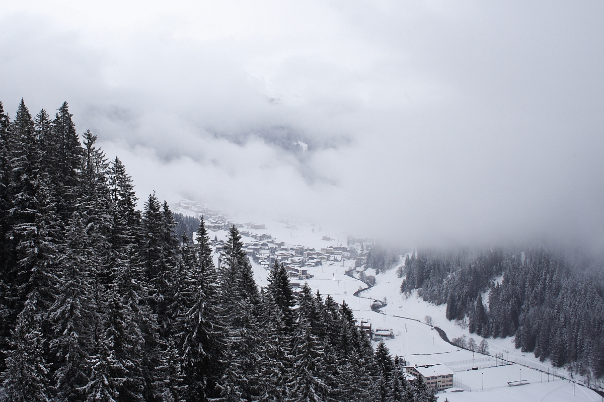 A snow-covered village nestled in a valley surrounded by dense pine forests and blanketed in mist and clouds lies within the Austrian Alps. Buildings are sprinkled throughout the landscape, and a winding road weaves through the snowy terrain. Tall trees in the foreground frame the serene, wintery scene.