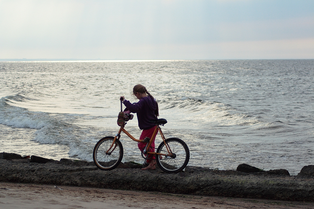 A young girl stands beside her bicycle on a rocky shore near the Daugavgrīva lighthouse, facing the ocean. She is wearing a purple hoodie and red pants. The ocean waves gently crash against the rocks, and the sky above is overcast with soft light filtering through the clouds. The beach stretches in the foreground.