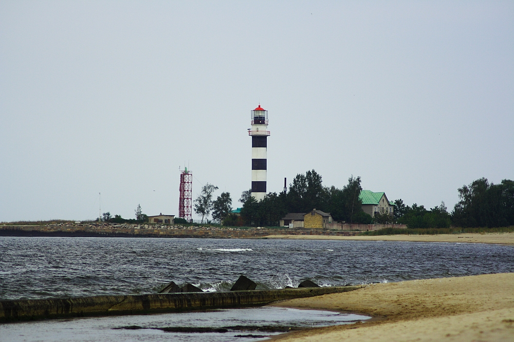 A coastal scene with the Daugavgrīva black and white striped lighthouse standing tall near the shoreline. Adjacent to the lighthouse are small buildings with green roofs. There is a red communication tower nearby. The beach has a gentle curve, with waves approaching the sandy shore under a cloudy sky.