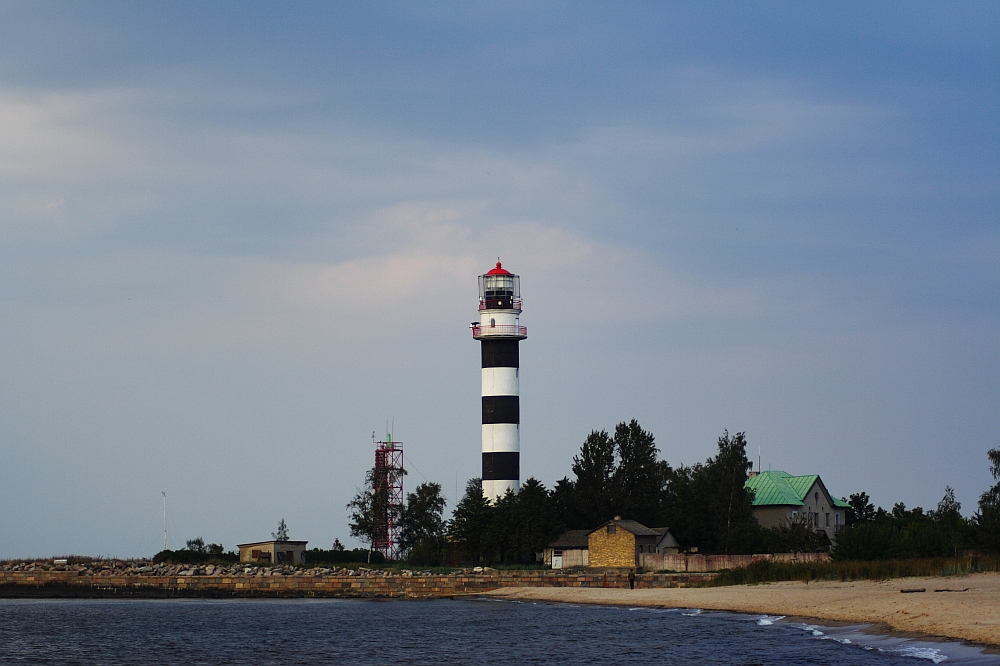 A black and white striped lighthouse stands on the coastal shore of Daugavgrīva under a cloudy sky. Surrounding the lighthouse are a few small buildings, including one with a green roof. The foreground has a sandy beach and calm ocean waves lapping the shore. Trees and a red and white tower are adjacent to the lighthouse.