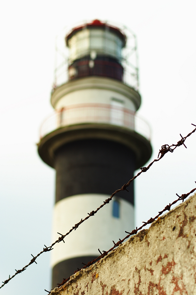 A tall lighthouse with black and white horizontal stripes stands slightly out of focus in the background. Known as the Daugavgrīva lighthouse, it features a lantern room at the top with protective railings. In the foreground, a rusty barb wire fence is sharply in focus, with a weathered concrete wall beneath it.