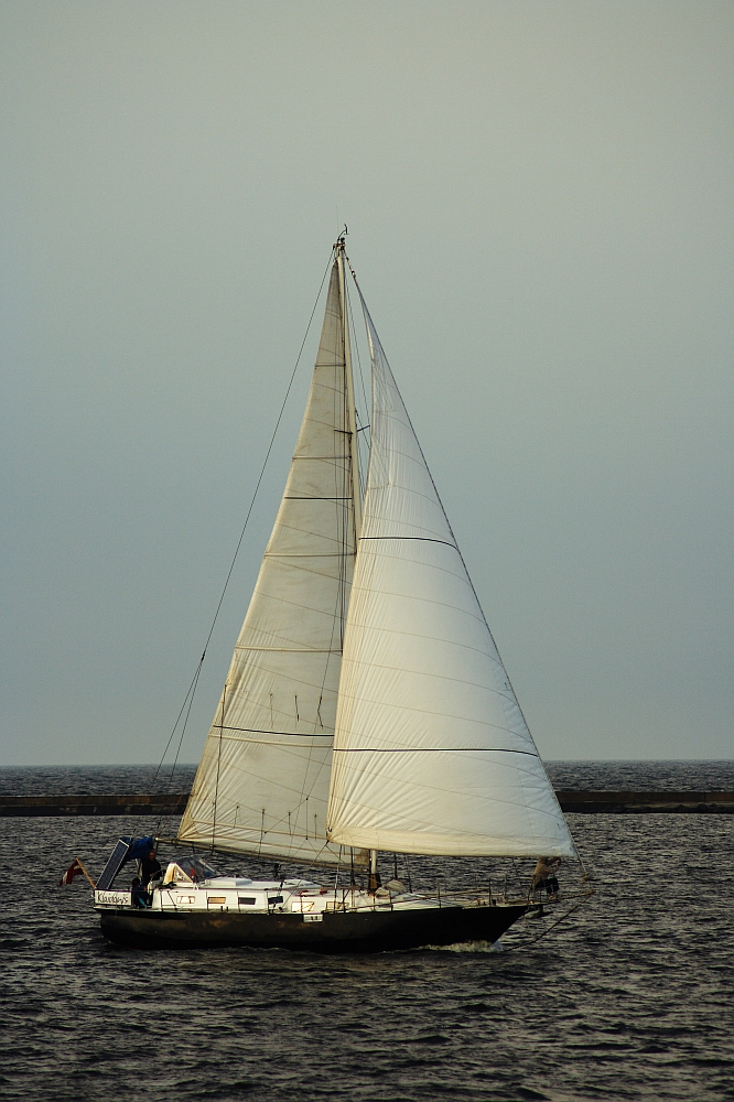 A lone sailboat with white sails fully extended glides on a calm body of water near the Daugavgrīva lighthouse. The sky above is overcast, and a distant, dark horizon line separates the sky from the water. Two people are on the boat's deck, possibly navigating or adjusting the sails.