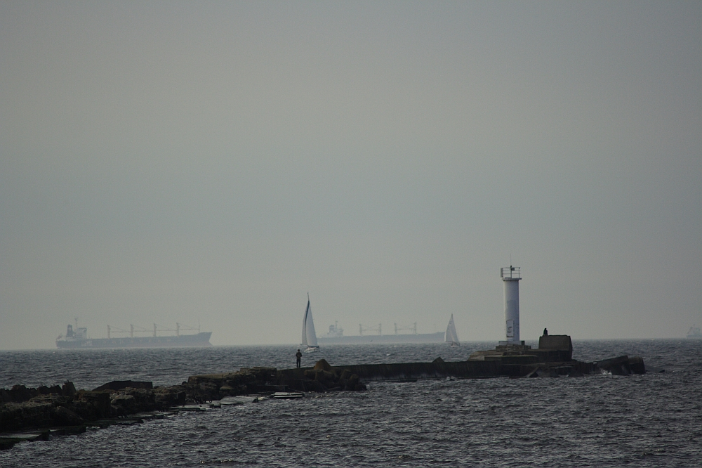 A hazy, overcast scene of a rocky shoreline extending into the sea with a small white lighthouse at Daugavgrīva and a few people near it. Several large ships and a sailboat are visible on the horizon under the gray sky. The water appears calm with gentle waves.
