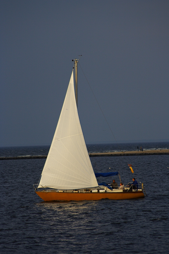 A single-mast sailboat with white sails and a yellow hull is sailing on calm blue waters near Daugavgrīva. Two people can be seen onboard under a blue canopy. The background features a narrow strip of land with a few people walking by the lighthouse, and the sky is a gradient from light to dark blue.
