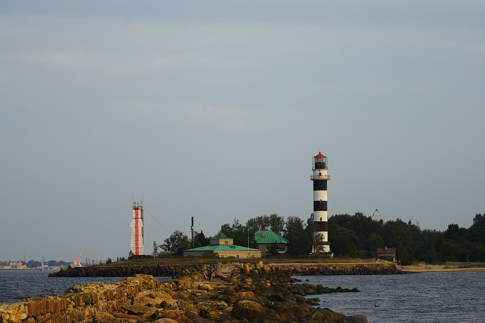 A rocky pier extends out into the water, leading to Daugavgrīva with two lighthouses. The near lighthouse is black and white, while the farther lighthouse is red and white. A small, green-roofed building stands between them, surrounded by trees. The sky is overcast.