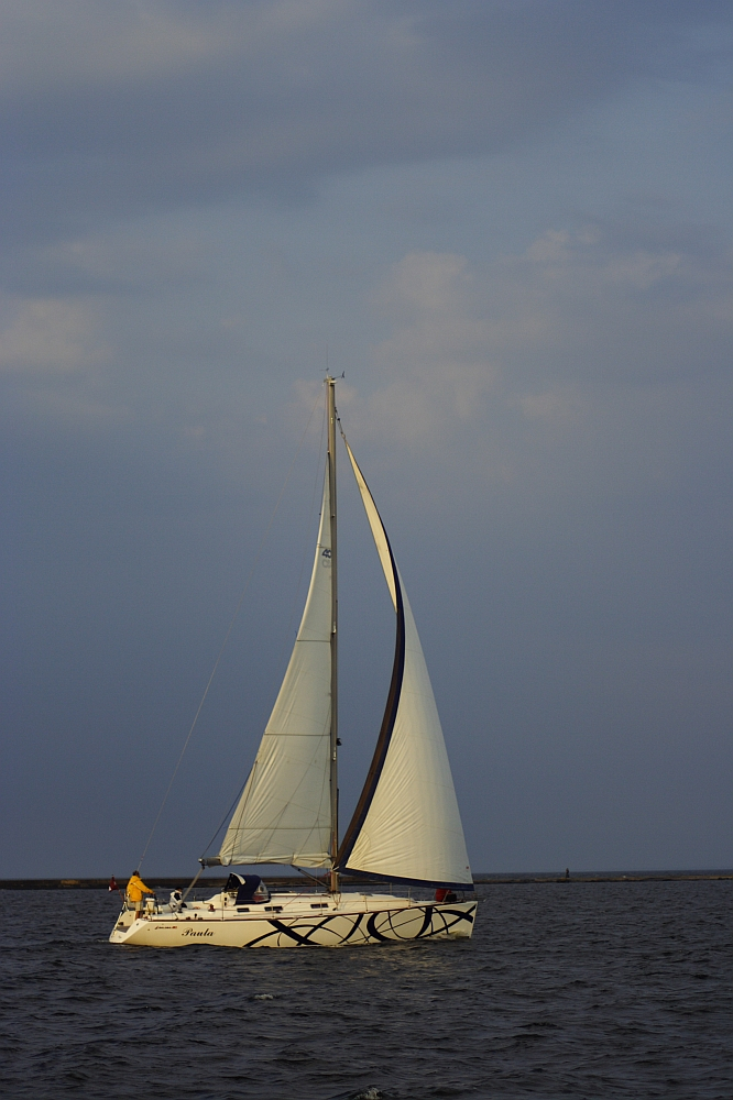 A white sailboat with intricate black designs on its hull glides through the water near the Daugavgrīva lighthouse. Two people are on board, one wearing a yellow jacket. The boat’s sail is fully set, catching the wind. The sky is overcast with shades of blue and gray, and the water appears calm.