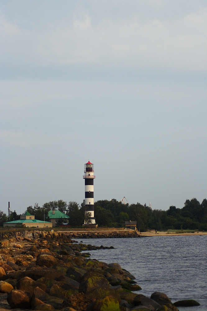 A striped lighthouse with black and white horizontal bands stands near a rocky shoreline, reminiscent of the Daugavgrīva lighthouse. Surrounding the scene are several small buildings and trees. The calm water of the sea extends towards the right, under a cloudy, light blue sky.