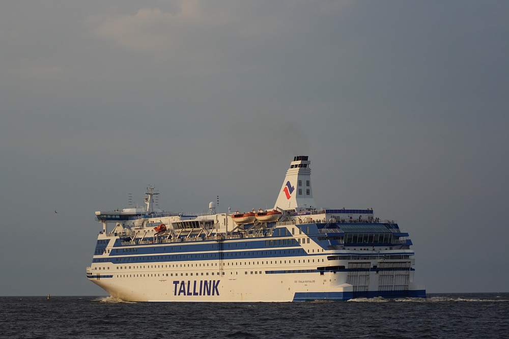 A large white passenger ferry with the name "TALLINK" in blue letters on the side sails past a distant lighthouse on the calm sea. The ship has several decks with visible lifeboats and is topped by a white smokestack with a blue and red logo. The sky above Daugavgrīva is mostly clear with a few clouds.