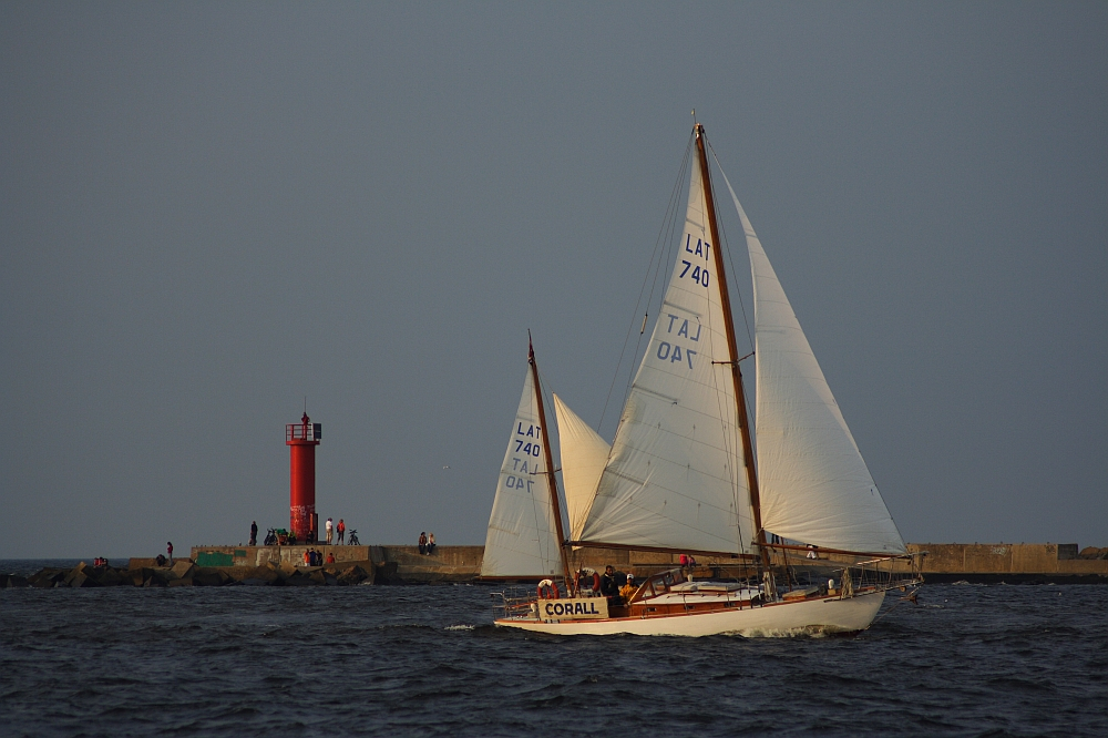 A sailboat named "Corall" with sail number LAT 740 glides on dark waters near a rocky breakwater. A vivid red lighthouse at Daugavgrīva stands on the breakwater, contrasting with the muted sky and sea. Several people are visible near the lighthouse, and the scene is calm, suggesting a peaceful day.