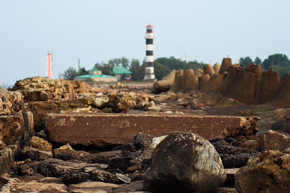 A rocky coastal area with large boulders in the foreground. Further back, the Daugavgrīva lighthouse stands tall with its black and white stripes, surrounded by green-roofed buildings and trees. The sky above is clear and slightly hazy, providing a calm atmosphere.