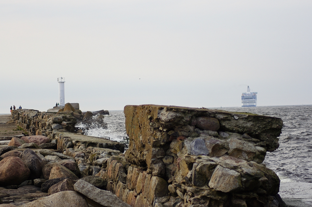 A stone pier extends into the sea, with large rocks scattered along its path. Near the end of the pier, Daugavgrīva lighthouse stands proudly. In the distance, a blue and white ferryboat sails on the horizon under an overcast sky. Two people are visible walking on the pier.