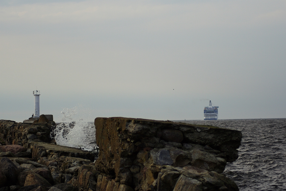 A rocky shoreline with large waves crashing against the rocks. A white lighthouse stands on the left side of the image near the edge of the shore, reminiscent of Daugavgrīva's historic beacon. In the distance, a large ferry can be seen on the horizon of the open sea under a cloudy, pale sky.