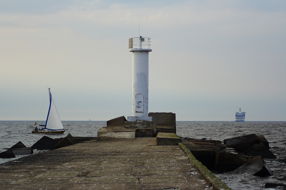 A solitary white Daugavgrīva lighthouse stands at the end of a weathered, concrete jetty extending into calm sea waters. To the left, a small sailboat with a white sail glides by, while a large ferry is visible in the distance on the right. The sky above is overcast with a pale, muted light.