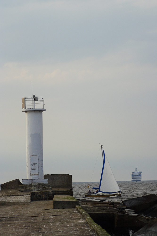 A white lighthouse stands at the edge of a concrete pier extending into the sea at Daugavgrīva. A sailboat with a white hull and blue sail is nearby in the water. In the distance, a large ferry or cruise ship is visible on the horizon under a cloudy sky. The scene exudes a calm, maritime atmosphere.