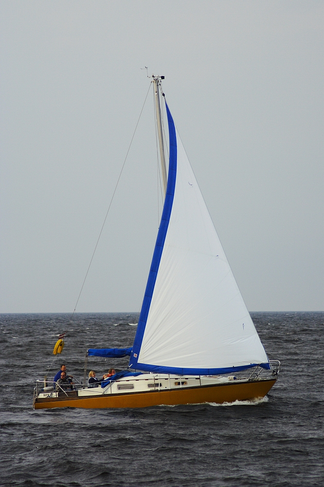 A yellow and white sailboat is sailing on a body of dark, choppy water, near the historic Daugavgrīva lighthouse. The sail is primarily white with a blue trim along the edges. There are a few people on board, wearing jackets. The sky is overcast, suggesting a cloudy day. A small yellow buoy is visible in the distance behind the sailboat.