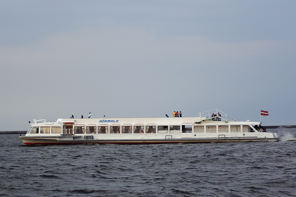 A white passenger ferry named "Jūrmala" is sailing on the waters of Daugavgrīva under a cloudy sky. The ferry, with its large windows, is flying a red and white flag at the stern. People on deck are enjoying the view, possibly spotting the distant lighthouse as they navigate slightly choppy waters.