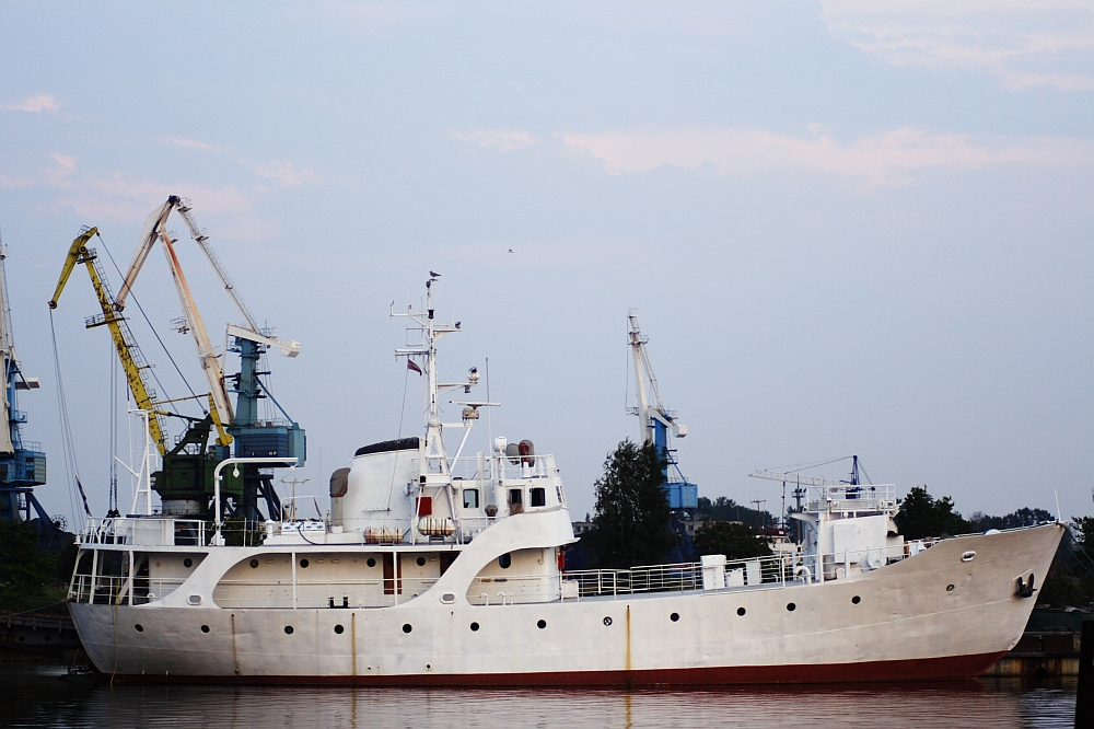 A vintage white ship docked at a harbor, featuring a rounded stern and multiple decks with various structures. Several cranes, painted yellow and blue, stand in the background near the Daugavgrīva lighthouse. The ship reflects a serene evening sky with a hint of pink, along with the calm water surface it is moored on.
