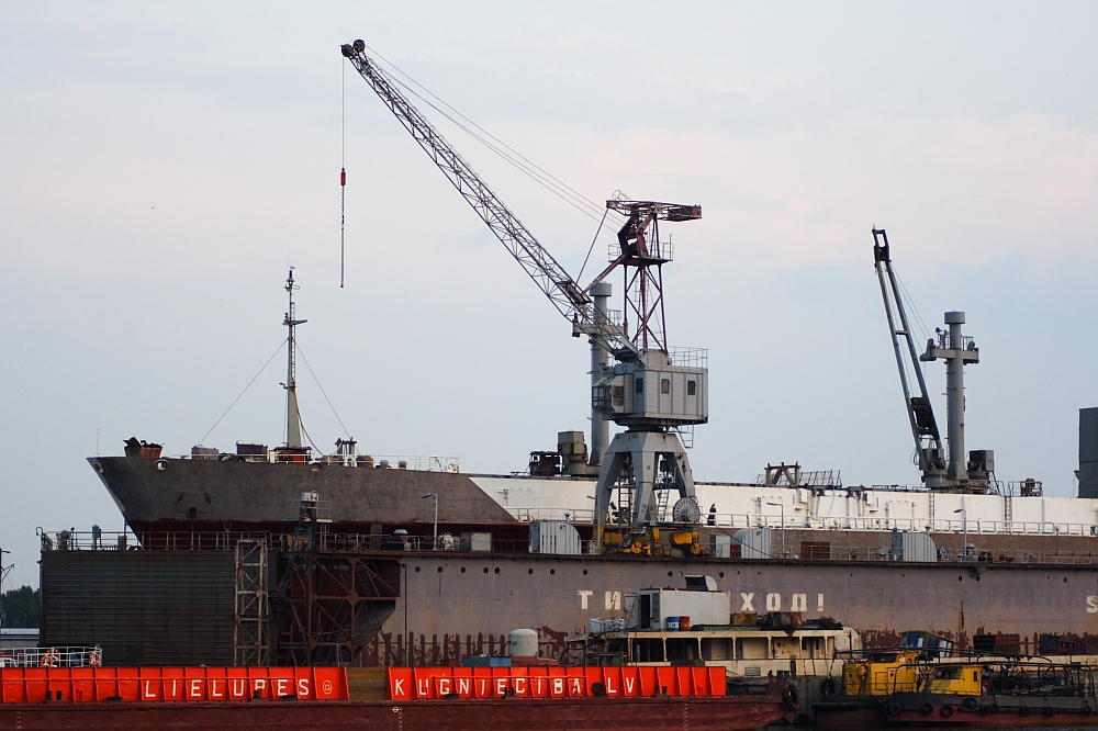A large ship is docked for repairs in a Daugavgrīva shipyard. Two cranes are visible: one in the foreground lifting equipment onto the ship and another in the background. The lower part of the hull displays weathering marks. Red barriers with unreadable white text can be seen, and a distant lighthouse stands sentinel.