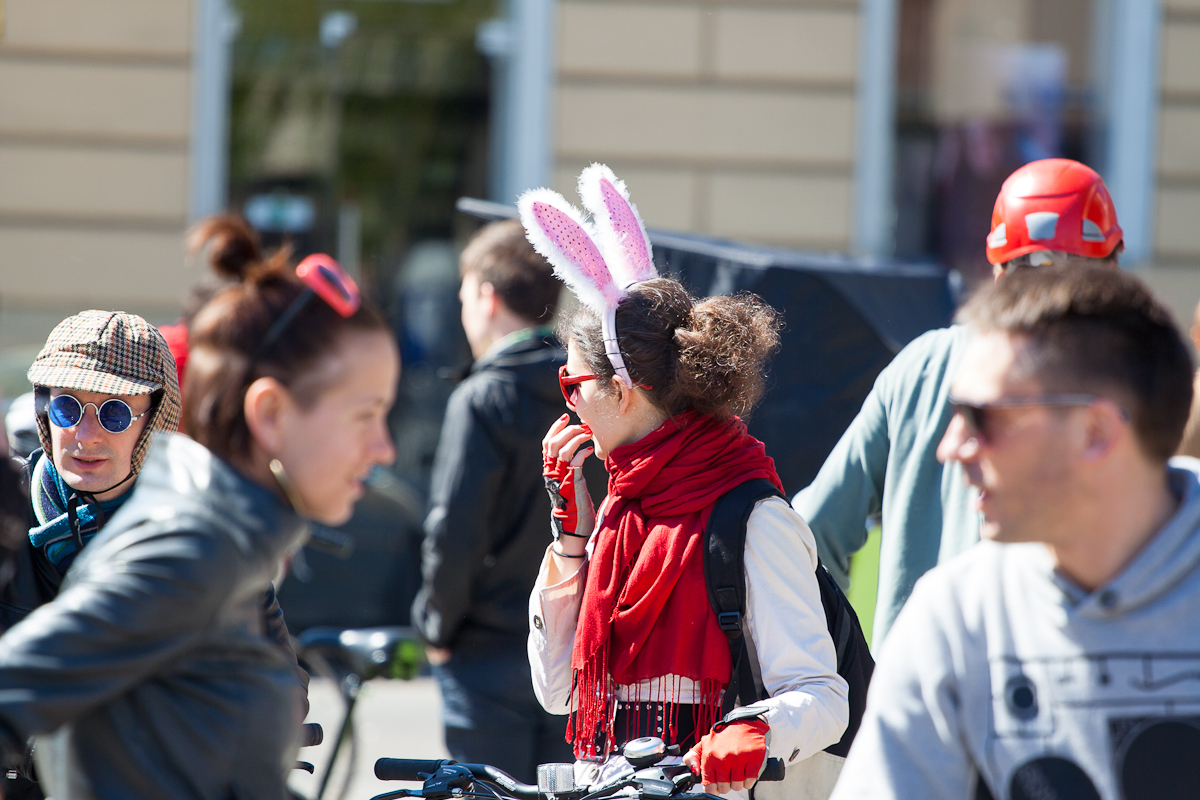 People are gathered outdoors in bright sunlight, some standing next to bicycles. The focus is on a person wearing white rabbit ears and a red scarf, reminiscent of Kritiskā masa 2012. Around this individual, others engage in conversation—one donned sunglasses and another a red helmet—with a blurred building in the background.