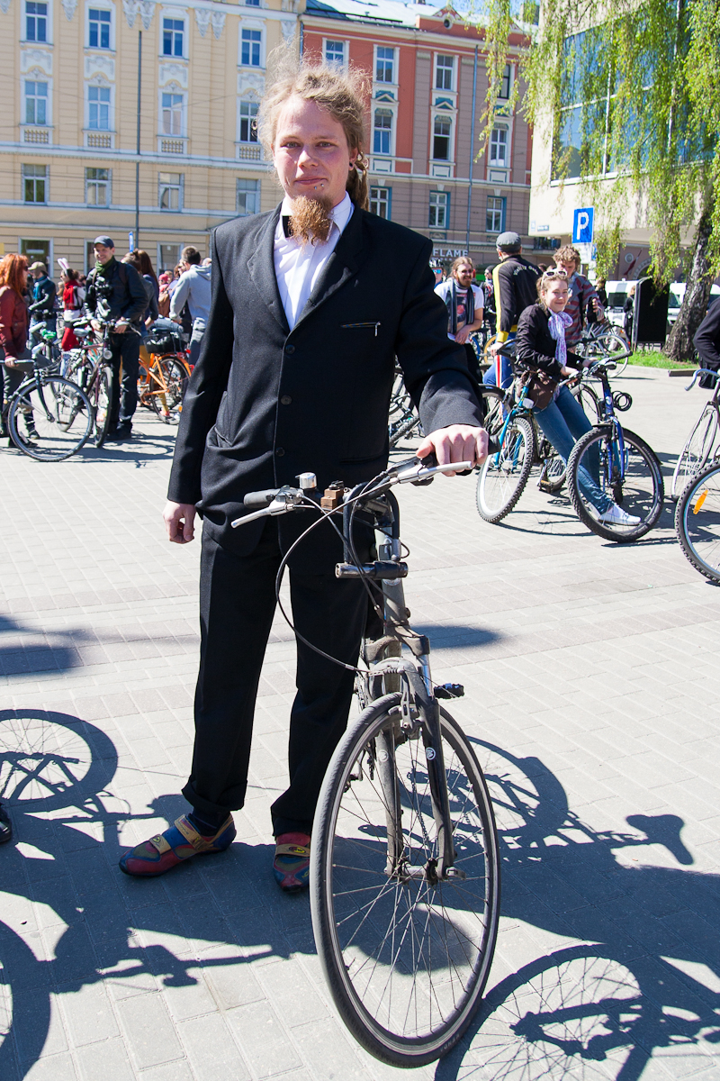 A man with a beard and mustache stands outdoors, holding a bicycle during the 2012 Kritiskā masa event. He is wearing a black suit with a white dress shirt and colorful shoes. Several other people with bicycles are in the background, with buildings and trees visible on this sunny day.