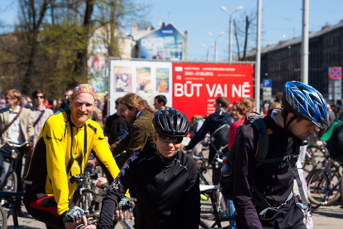 A group of cyclists gathered in an urban area, mostly wearing helmets and sports attire. One cyclist in a yellow jacket stands out, while others blend in with darker clothing. Behind them is a large red billboard with white text and surrounding people, buildings, and trees in the background from Kritiskā masa 2012.