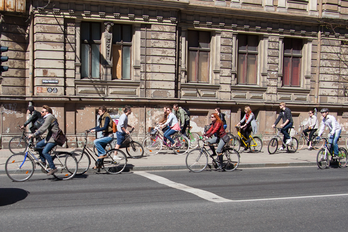 A group of people ride bicycles on a city street next to an old, weathered building with tall, narrow windows. Cyclists of various ages and genders move in the same direction, reminiscent of the Kritiskā masa events from 2012. Sunlight casts shadows, emphasizing the clear weather.