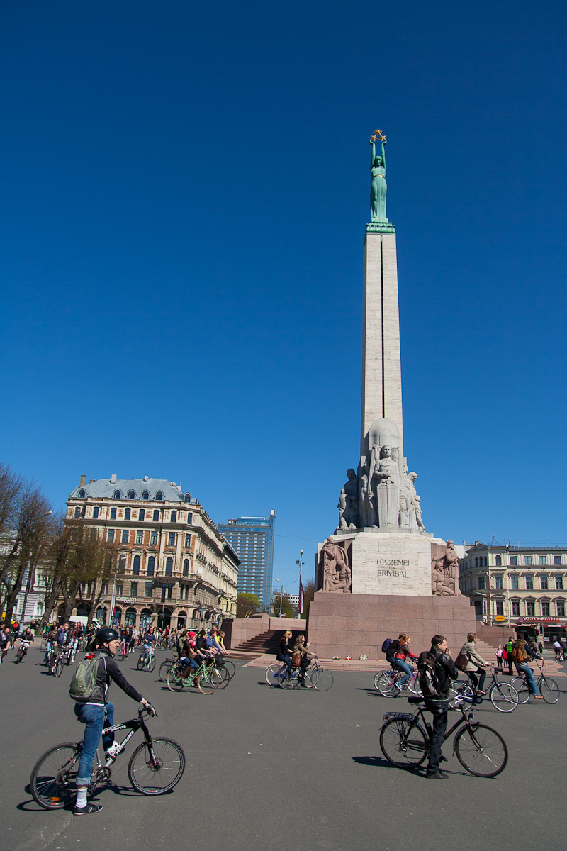 A tall monument with a statue holding three stars stands in an urban square under a clear blue sky. Cyclists and pedestrians, reminiscent of the 2012 Kritiskā masa movement, are actively moving around the square. Historic buildings with ornate architecture are visible in the background, along with a modern high-rise.