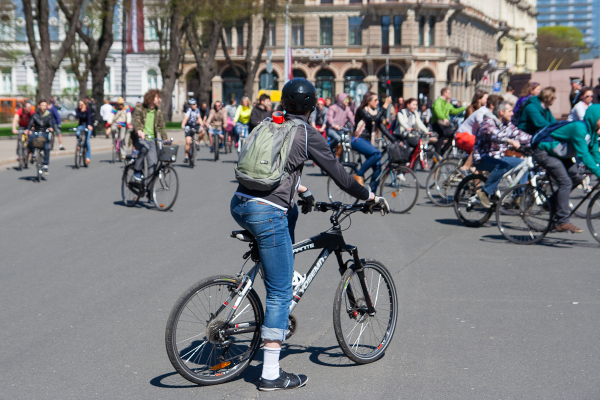 A group of cyclists ride on a city street during Kritiskā masa in Riga, a public event held in 2012. The foreground features a person wearing a black helmet, green backpack, black jacket, and blue jeans on a black bike. Dozens of other cyclists, pedestrians, and historic buildings are visible in the background. The weather is sunny.