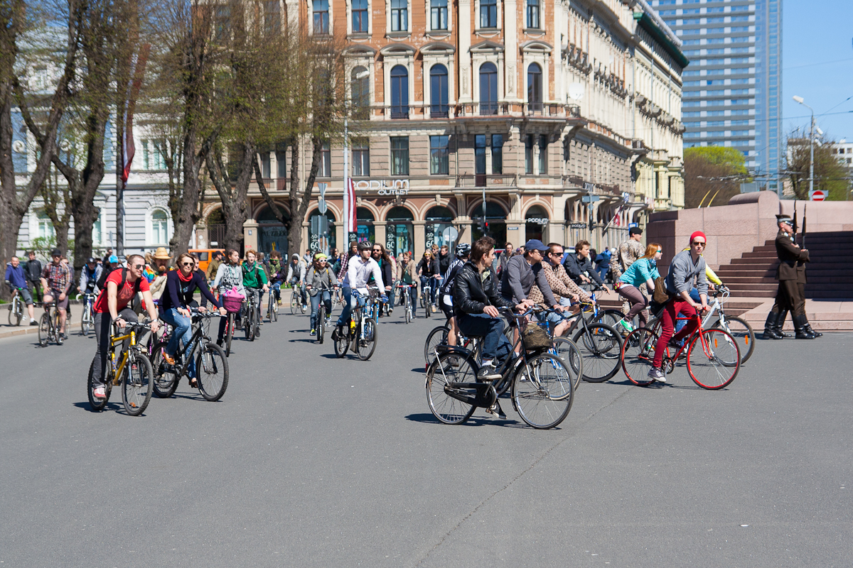 A large group of people are riding bicycles on a wide street in an urban area with classic and modern buildings, reminiscent of the 2012 Kritiskā masa. Some cyclists wear helmets and vibrant clothing. Trees with budding leaves line the street. Pedestrians walk on the sidewalks in the background on a sunny day.