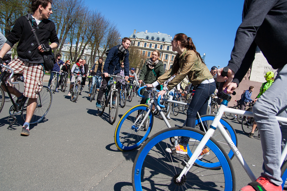 A group of people are participating in a cycling event on a sunny day, reminiscent of Kritiskā masa 2012. They are riding bicycles of various colors and styles. Trees with bare branches and a large, classic architectural building are visible in the background. Some cyclists appear to be conversing while riding.