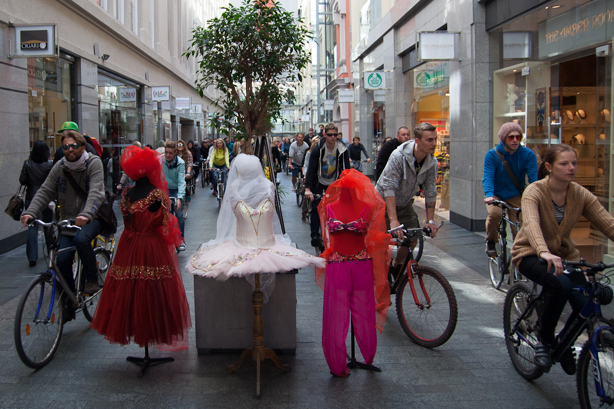 A busy shopping street with people walking and riding bicycles in both directions. Three mannequins dressed in vibrant clothes are displayed on a central pedestal: a white dress, a red dress, and a red outfit with a headscarf. Behind them stands a large, leafy plant. It feels almost like Kritiskā masa 2012 set against this vibrant urban backdrop.