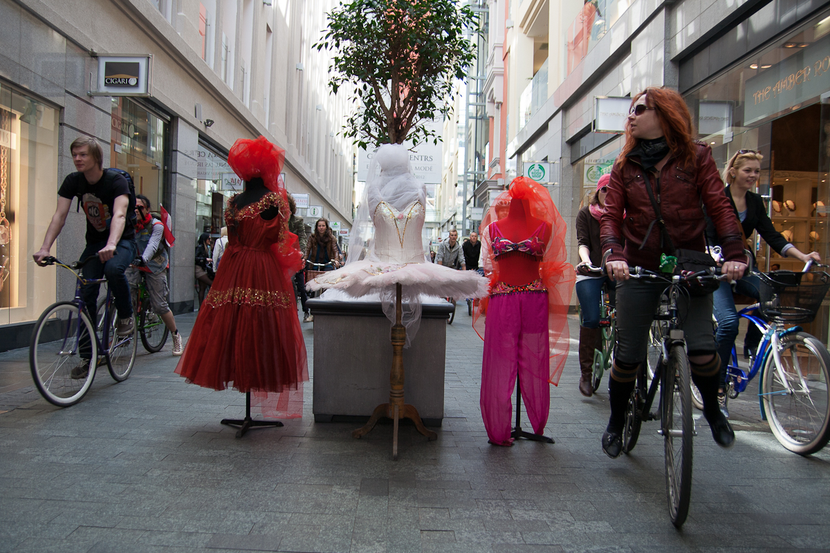 A street scene with cyclists from the 2012 Kritiskā masa passing by three mannequins on display. Two mannequins wear red dresses and veils, while the center one showcases a white tutu. The cyclists, a mix of men and women, ride through the shopping alley flanked by store windows and a potted tree.
