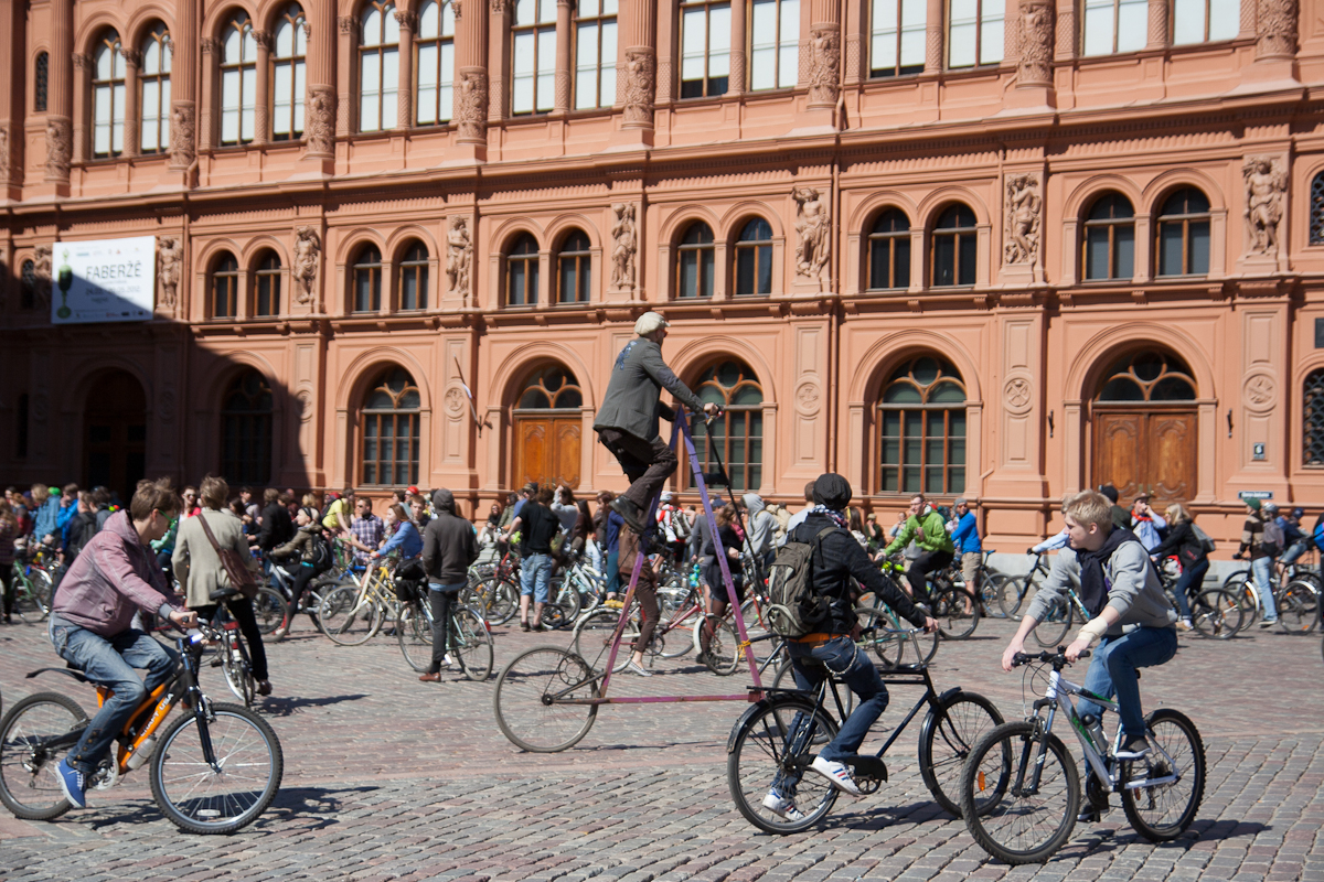 A lively scene unfolds in front of a large, ornate red-brick building with arched windows as a crowd of people participate in the 2012 Kritiskā masa event. Cyclists chat and ride, including one man on an unconventional tall bicycle towering above the rest, all under the sunlit sky on cobblestone pavement.