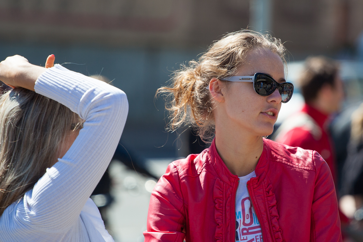Two women are in conversation outdoors. One woman, wearing dark sunglasses and a red jacket, looks to the side. Her hair is tied back, and she is wearing a white top underneath the jacket. The other woman, with blonde hair, is raising her right hand, obscuring part of her face as if participating in Kritiskā masa 2012.