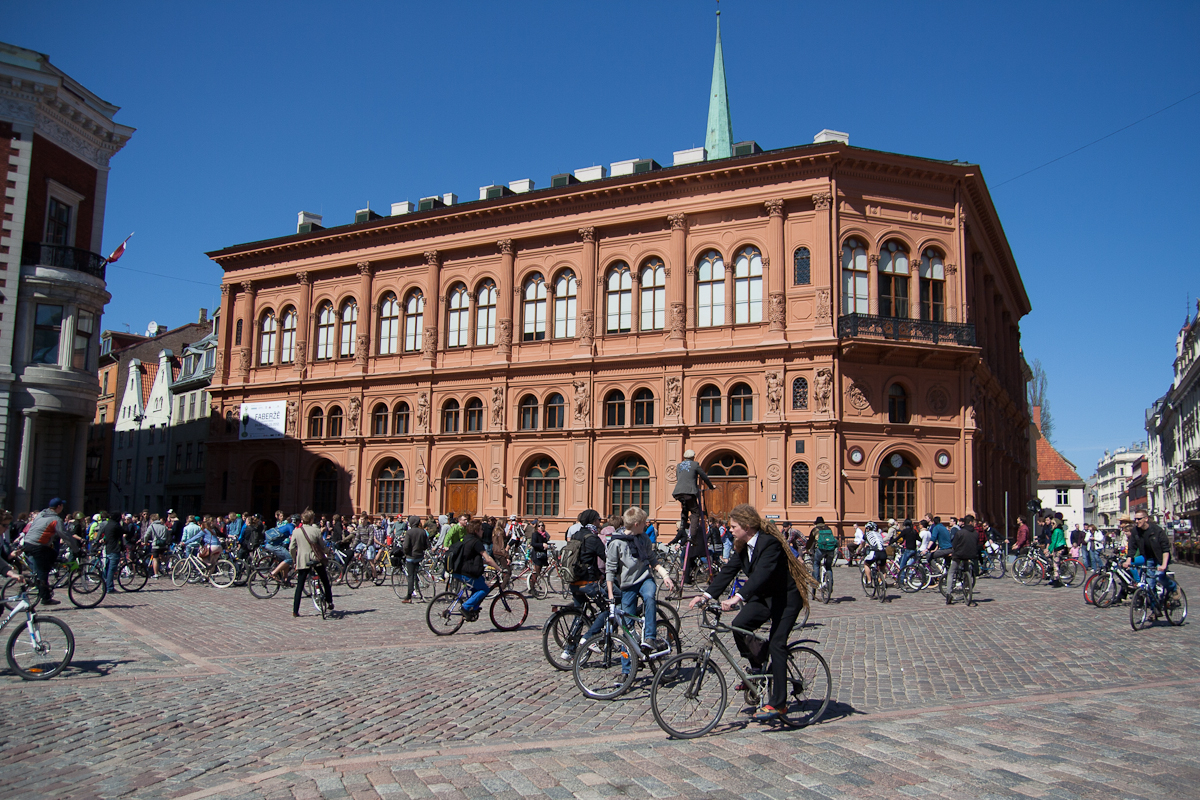 A bustling, sunlit square filled with cyclists participating in Kritiskā masa on cobblestone streets. A large, ornate terracotta-colored building with arched windows and a green spire dominates the background. Bright blue sky overhead, with neighboring buildings partially visible on the edges of the image—2012.