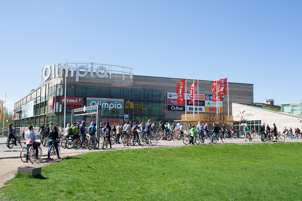 A busy scene in front of the large shopping center "Oulun Olympia" with multiple store signs. Numerous people are gathered, many with bicycles, perhaps participating in Kritiskā masa 2012, enjoying a bright, sunny day. Red flags and green grass add color while modern buildings stand under a clear blue sky.
